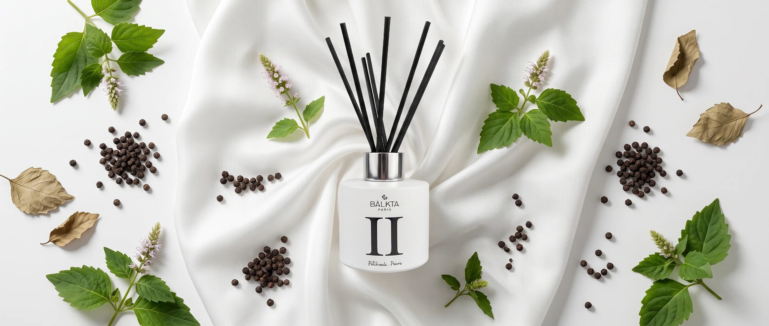 Diffuseur de parfum d'ambiance à bâtonnets BÂLKTA Numéro II (Patchouli & Poivre) entouré d'ingrédients frais et secs sur tissu de soie blanche, vue de dessus.