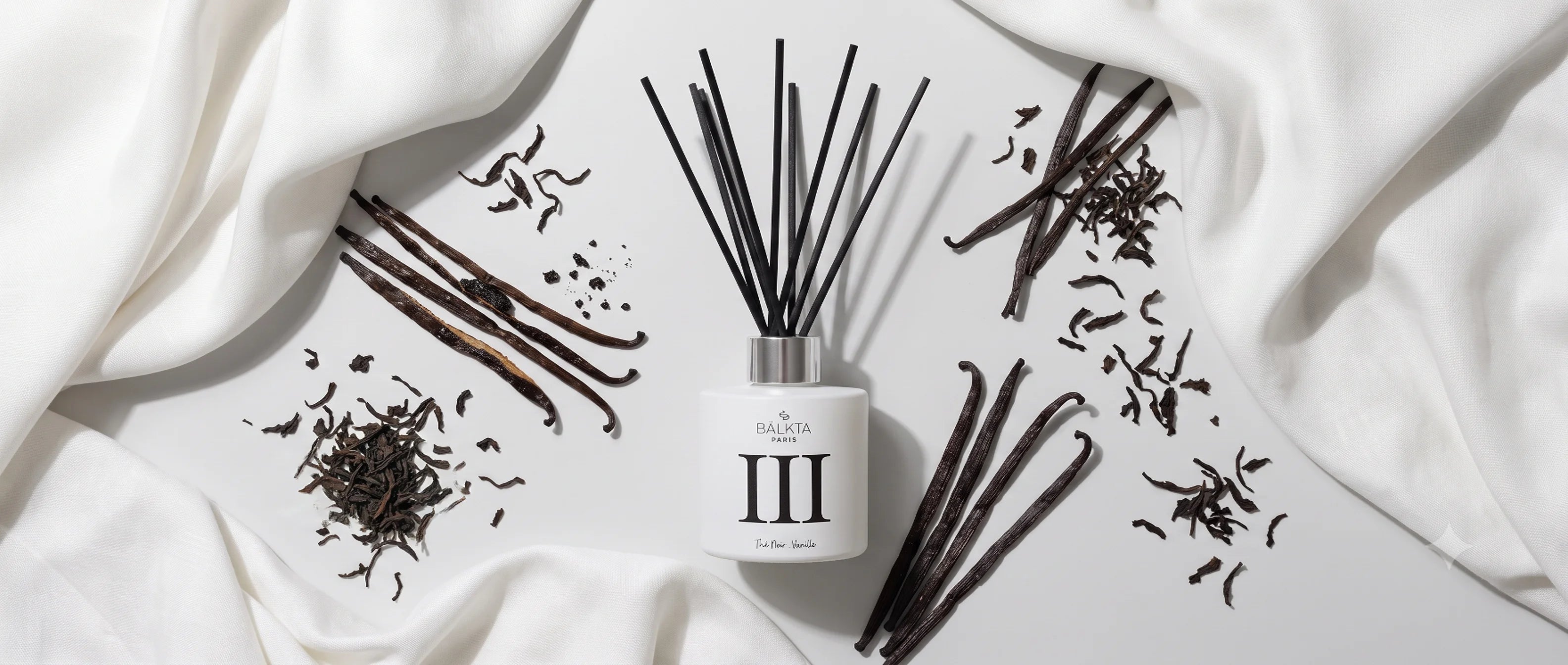 Diffuseur de parfum d'ambiance à roseaux Balkta Paris n°3 (Thé Noir - Vanille). Vue de dessus sur une surface claire entourée de gousses de vanille séchées, de feuilles de thé noir en vrac et de tissu en soie blanche.