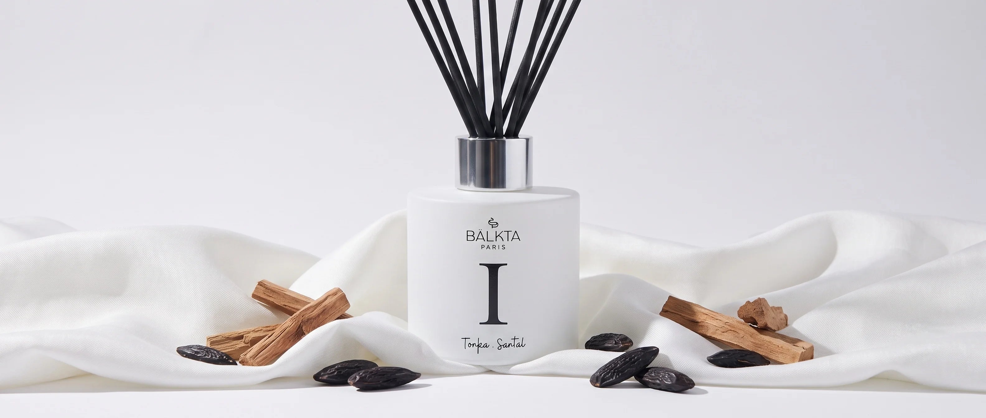 Diffuseur de parfum d'intérieur blanc à bâtonnets numéro I, mis en valeur avec des fèves tonka entières et des morceaux de bois de santal sur un élégant drapé blanc.