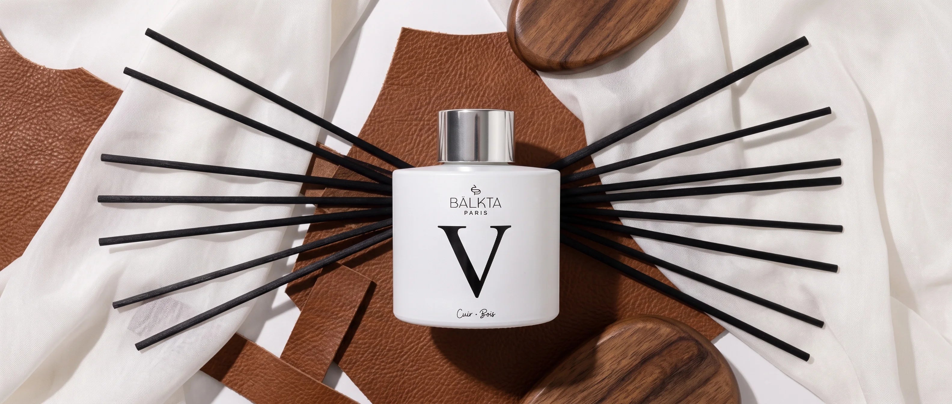 Diffuseur de parfum d'intérieur à bâtonnets Bälkta Paris numéro V, senteur Cuir et Bois, mis en scène sur des éléments en cuir et en bois avec un élégant drapé blanc.