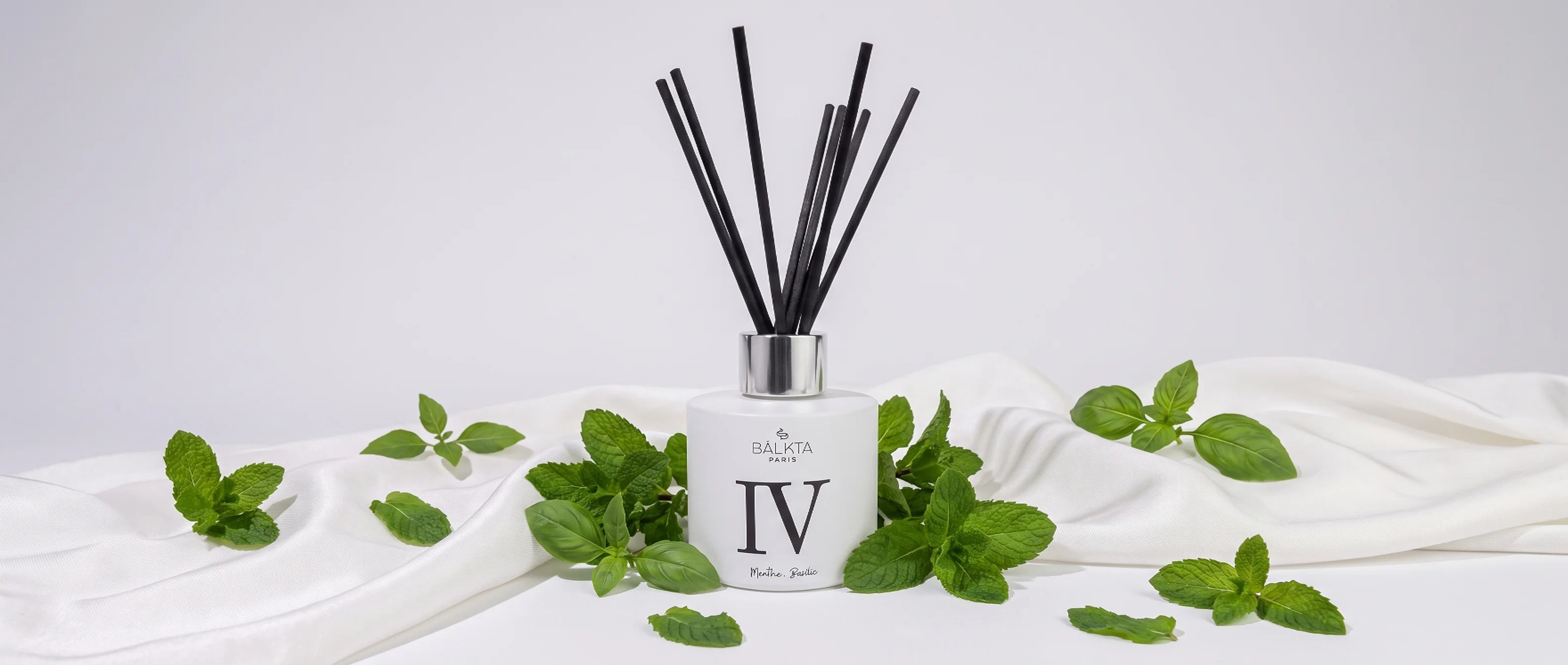 Diffuseur de parfum d'intérieur à bâtonnets Balkta Paris numéro IV, senteur Menthe et Basilic, entouré de feuilles fraîches sur un tissu blanc.