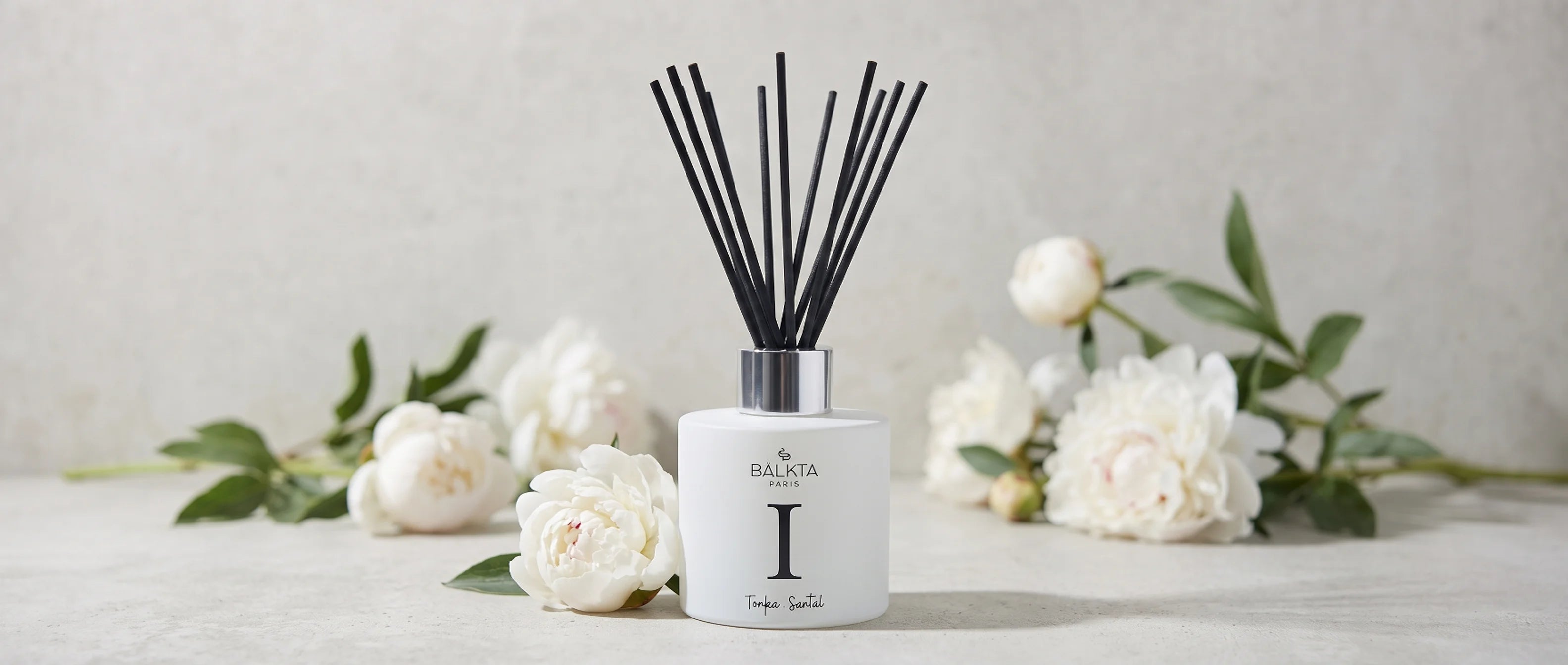 Diffuseur de parfum d'intérieur à bâtonnets Bälkta Paris numéro I, senteur Tonka et Santal, élégamment mis en scène avec des fleurs blanches sur un fond minéral clair.